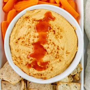 closeup of hummus drizzled with buffalo sauce and paprika surround with carrots and crackers