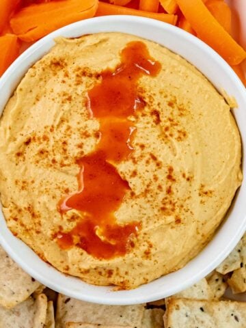 closeup of hummus drizzled with buffalo sauce and paprika surround with carrots and crackers