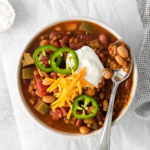 closeup of a white bowl full of lentil chili topped cheese, sour cream and three jalapeno slices with a silver spoon on the side