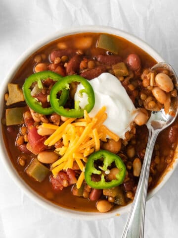 closeup of a white bowl full of lentil chili topped cheese, sour cream and three jalapeno slices with a silver spoon on the side