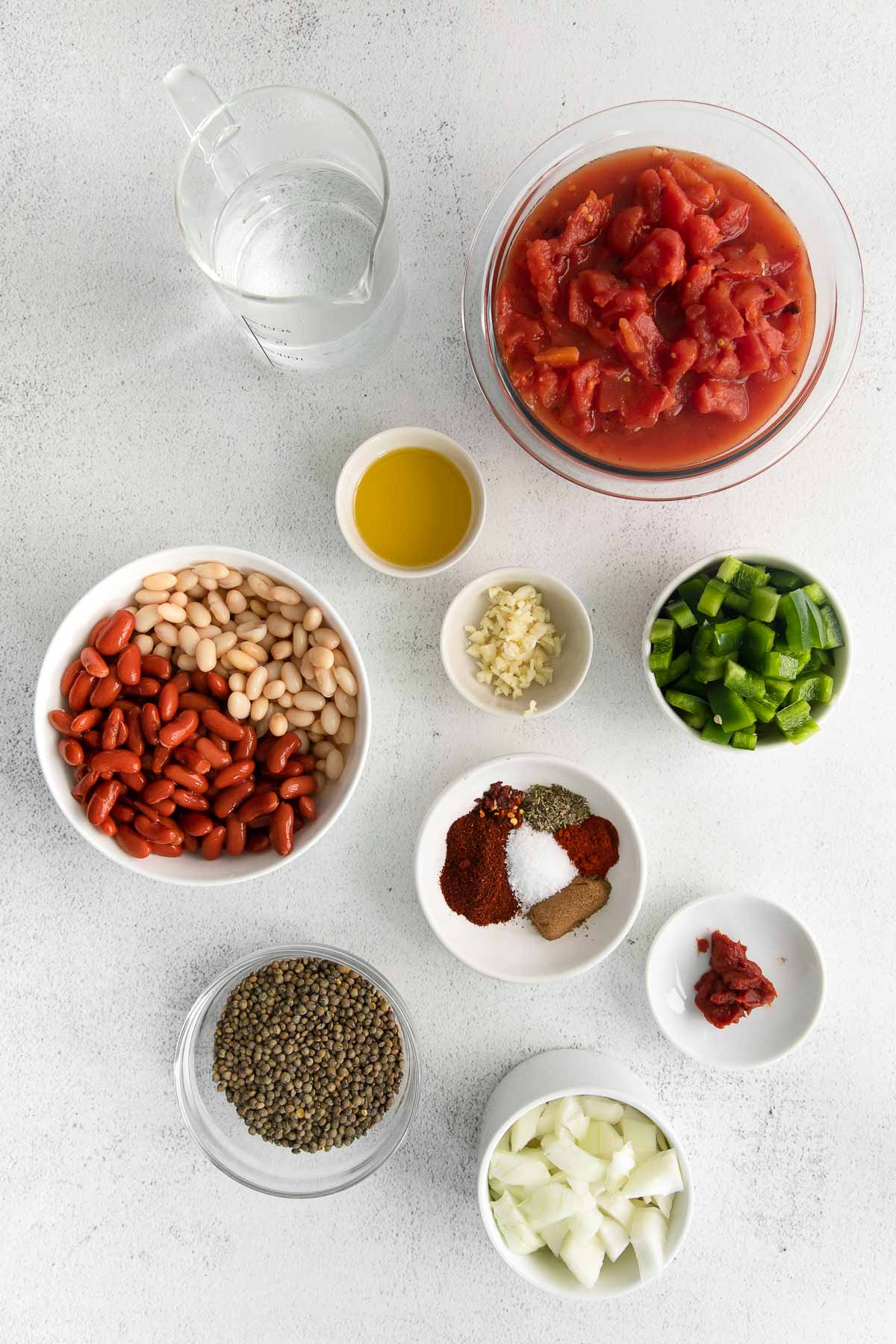 several small white and glass bowls full of ingredients for lentil chili - beans, diced onion, lentils, diced tomatoes, spices, garlic, and diced green peppers