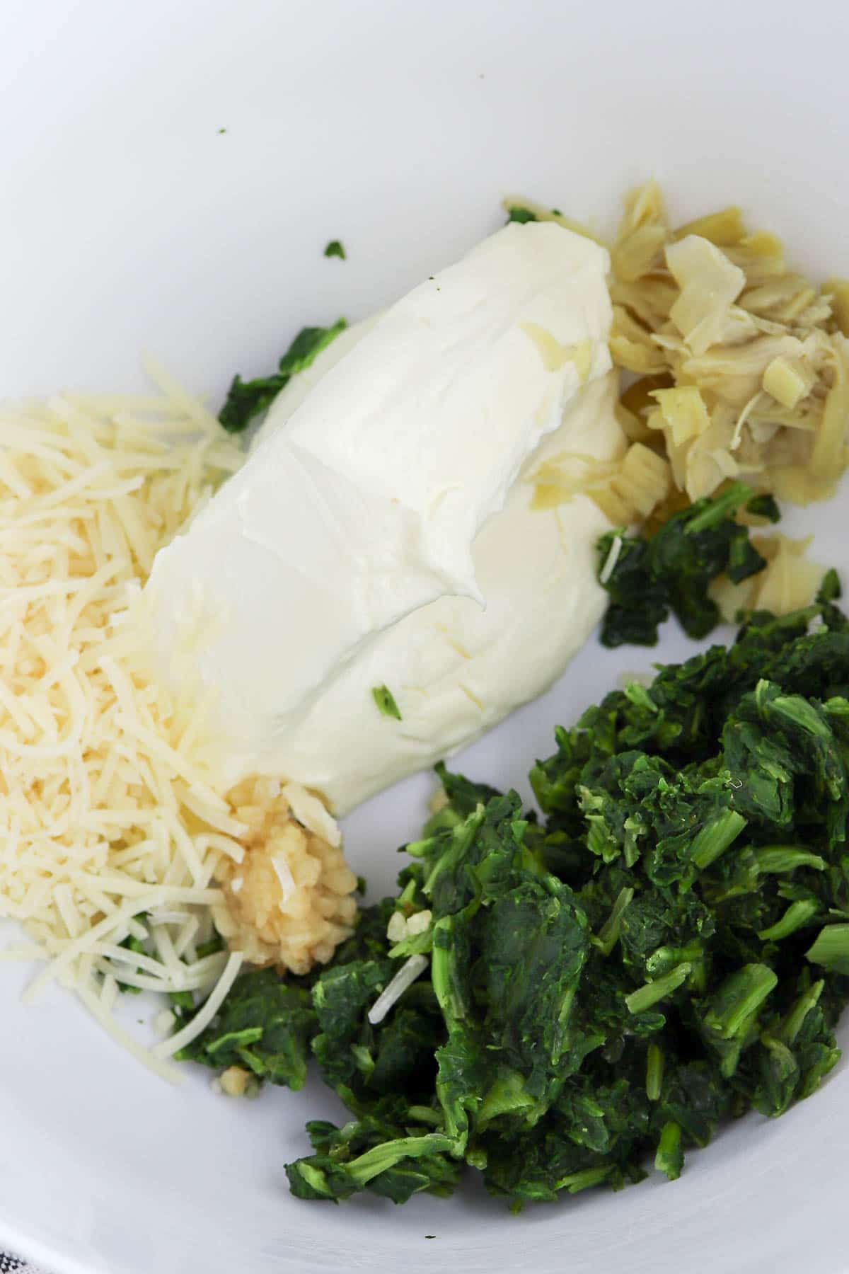 closeup of a white bowl filled with frozen spinach, chopped artichokes, parmesan cheese, cream cheese and minced garlic