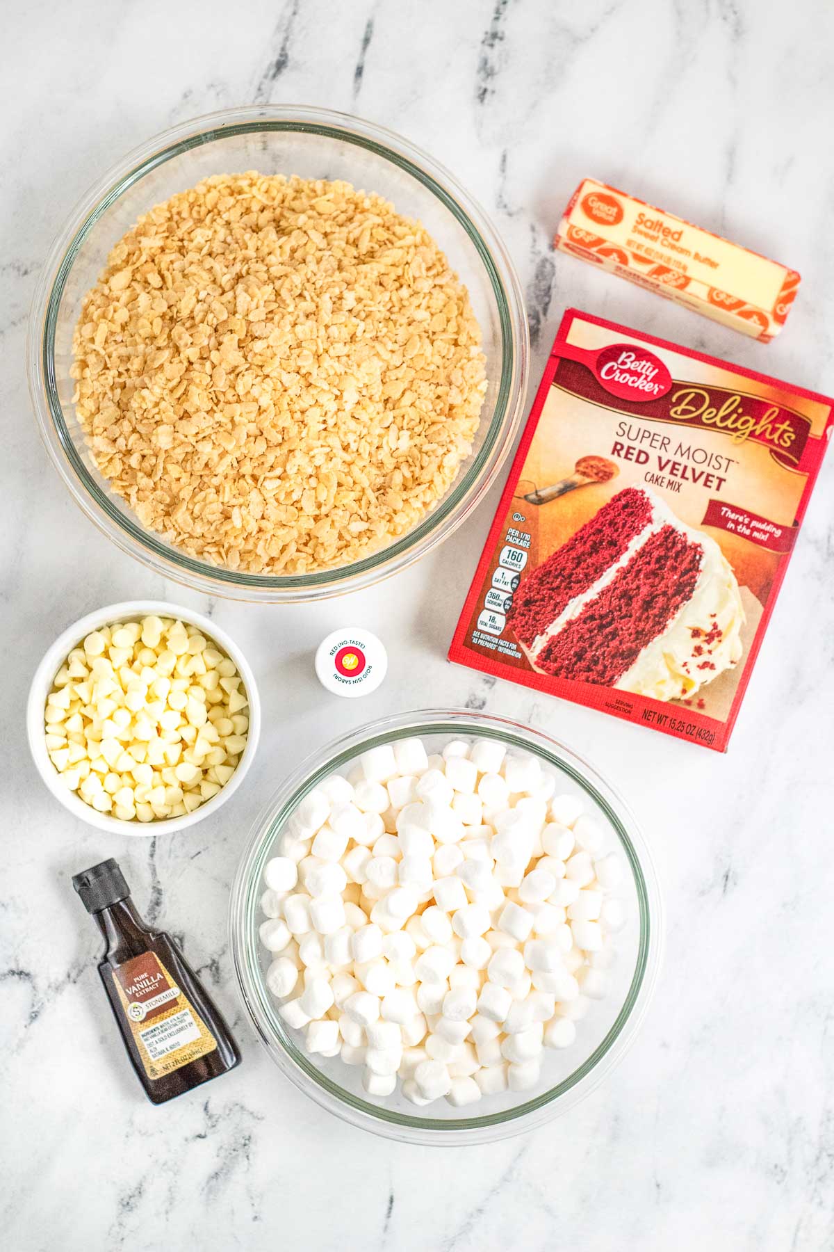 three bowls of mini marshmallows, rice krispie cereal, white chocolate chips, a box of red velvet cake mix, vanilla extract and a stick of butter