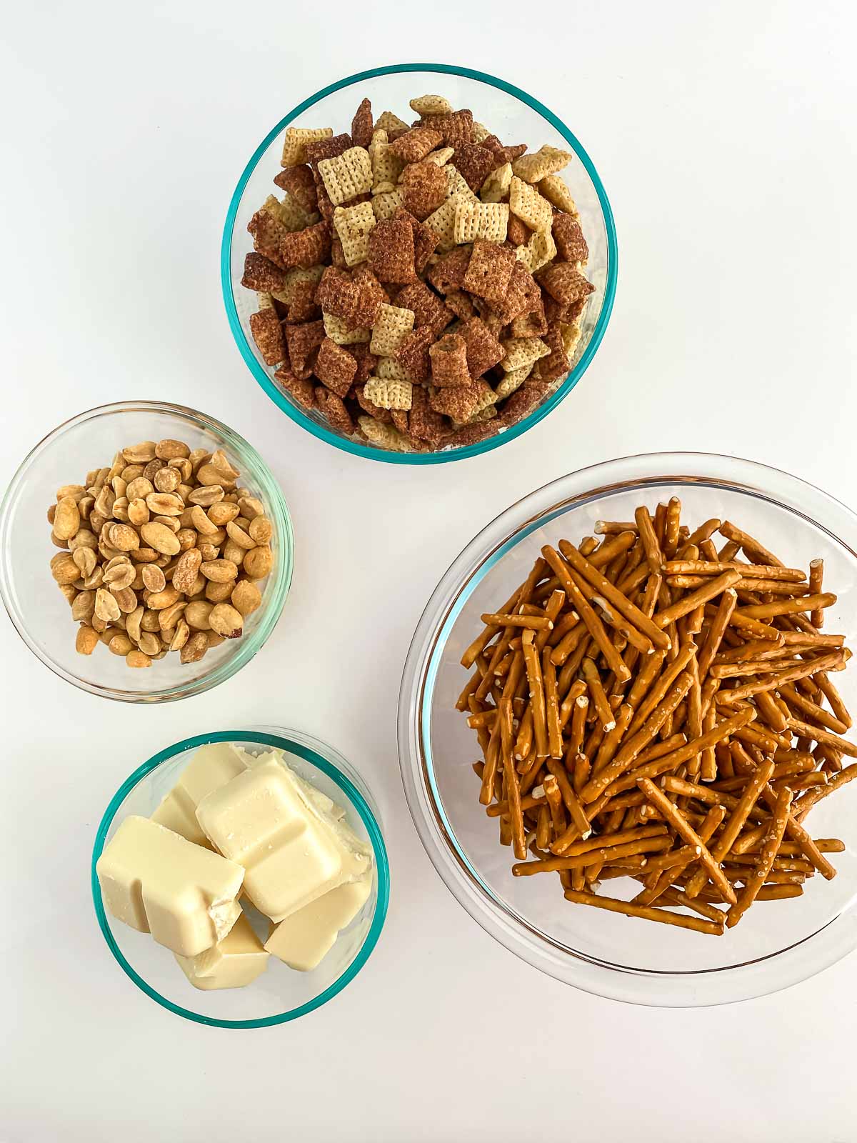 four glass bowls with pretzel sticks, chex cereal, peanuts and white chocolate squares
