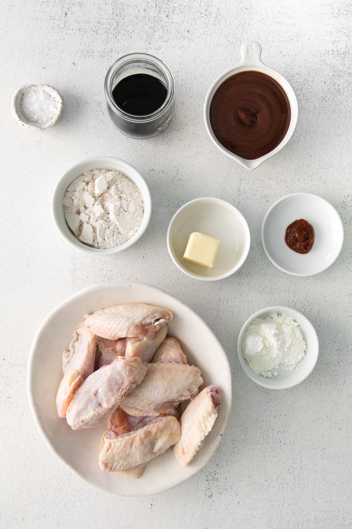 several white bowls with ingredients for baked chicken wings with bbq sauce mixture - raw chicken wings, bbq sauce, soy sauce, flour, corn starch, butter