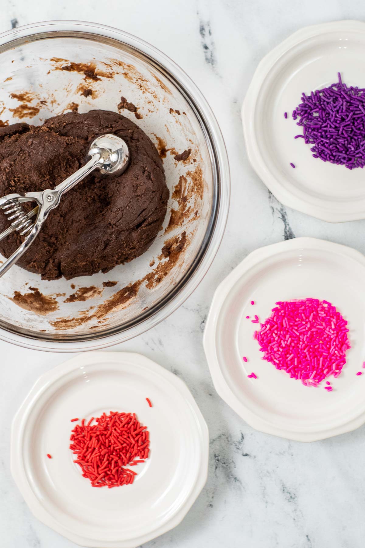glass bowl of brownie batter with a cookie scoop surrouded by three plates with pink, red and purple sprinkles