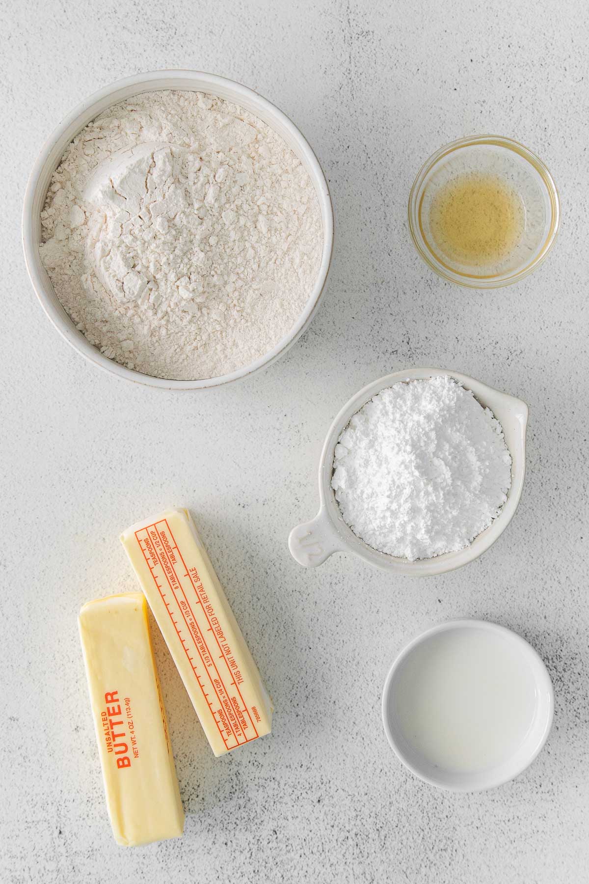 several white bowls of ingredients for shortbread cookies - milk, flour, powdered sugar and vanilla extract and two sticks of butter