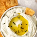 shallow bowl full of creamy feta dip topped with olive oil and fresh herbs and surrounds by pita chip crackers