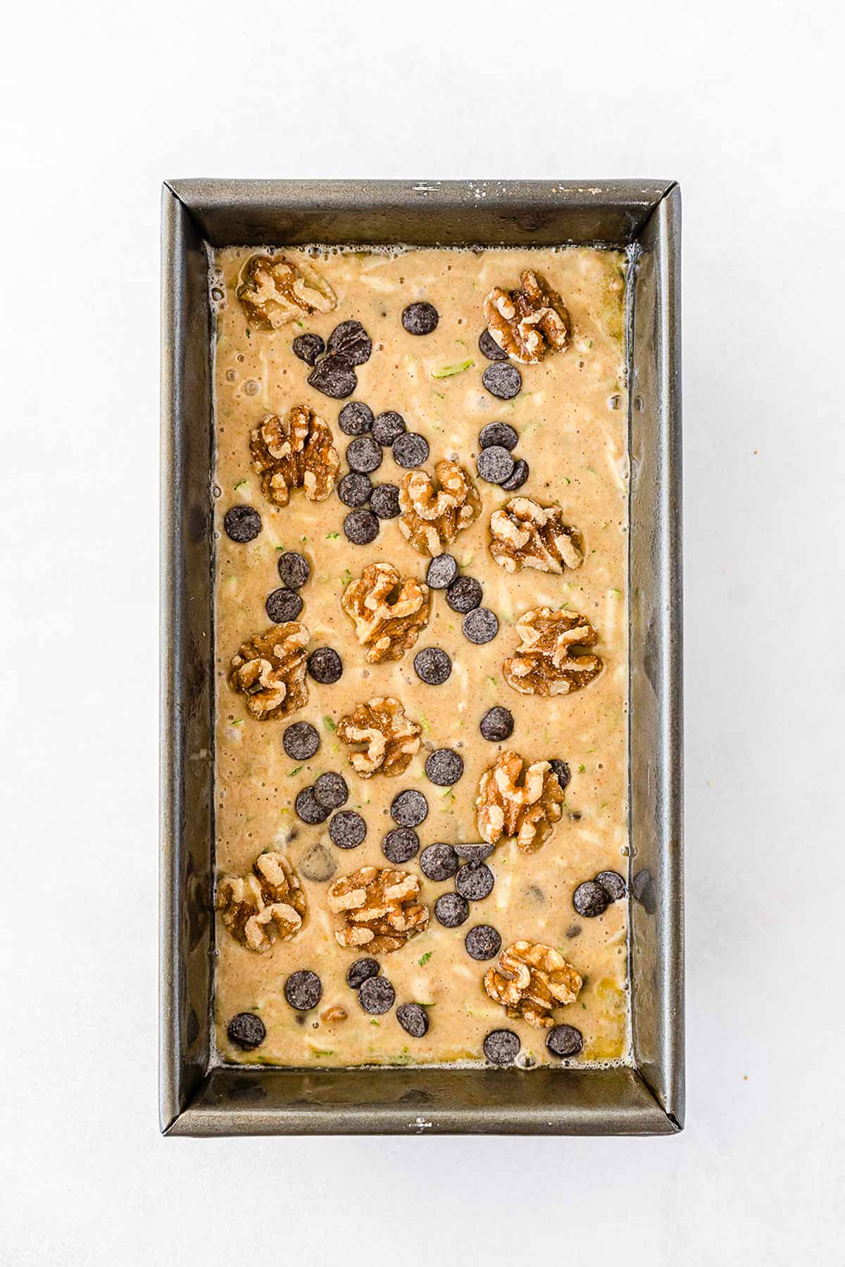 zucchini bread batter topped with chocolate chips and chopped walnuts in a silver loaf pan