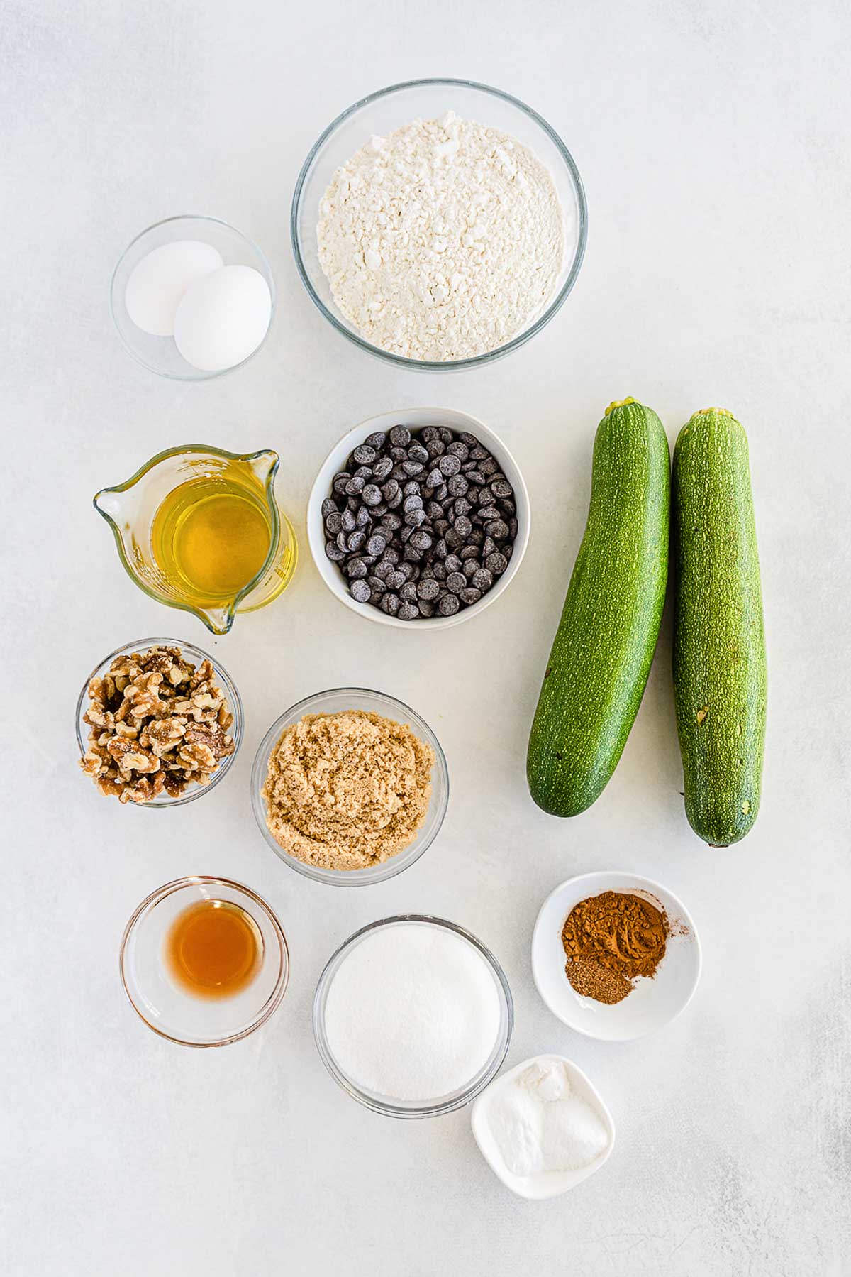 several glass bowls filled with ingredients for zucchini bread - two whole zucchini, brown sugar, white sugar, flour, cinnamon, baking soda, oil, chocolate chips and walnuts