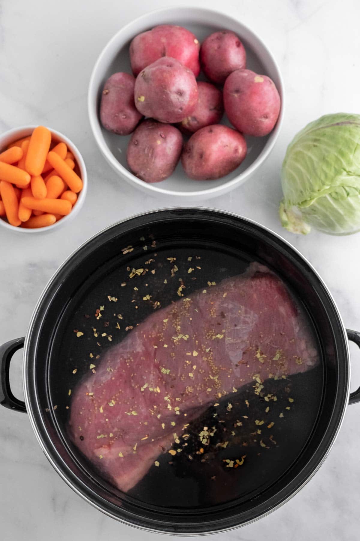 big black pot with a raw corned beef, a bowl of whole red potatoes, bowl of baby carrots and a head of cabbage