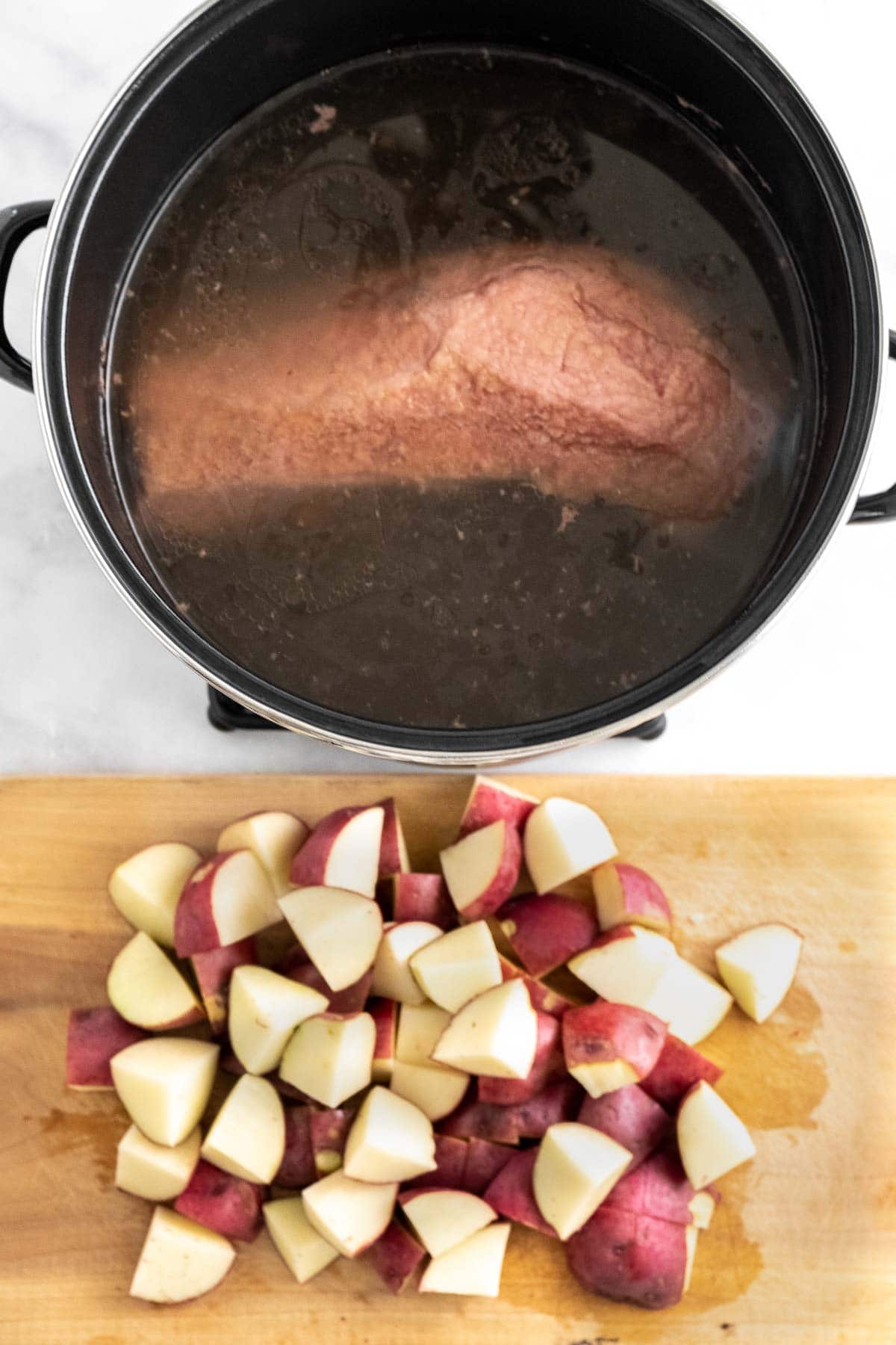 big black pot with cooked corned beef in water with a wood cutting board full of cut red potatoes