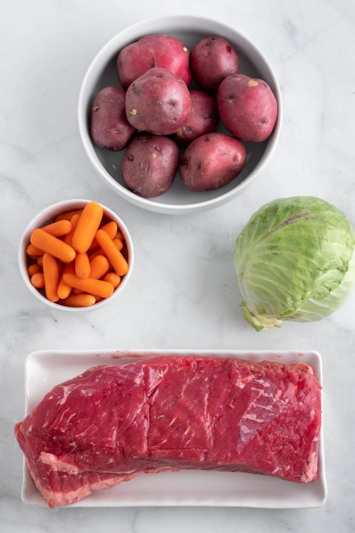 raw corned beef, bowl of red potatoes, bowl of carrots and a head of cabbage