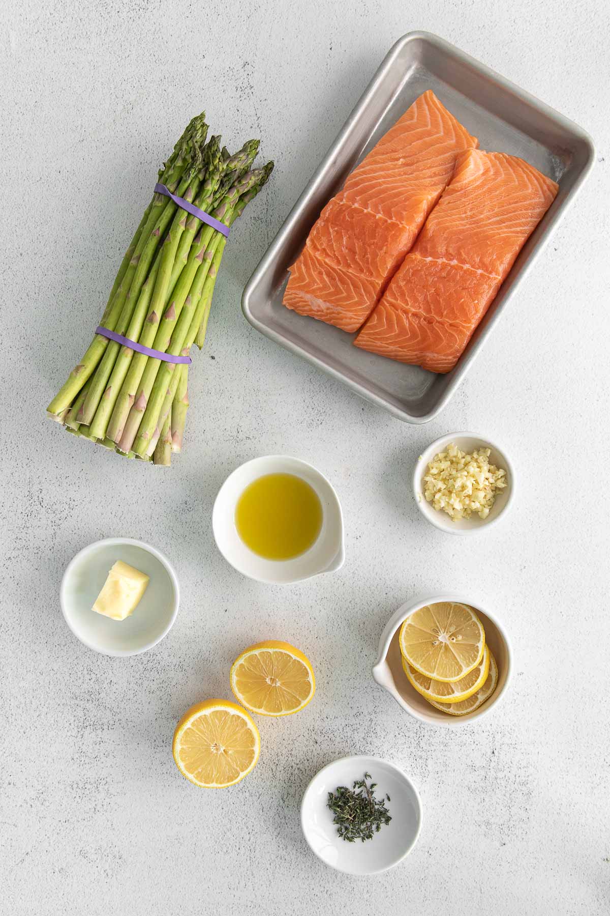 baking sheet with two salmon fillets, one bunch of asparagus, and several small white bowls with lemon, spices