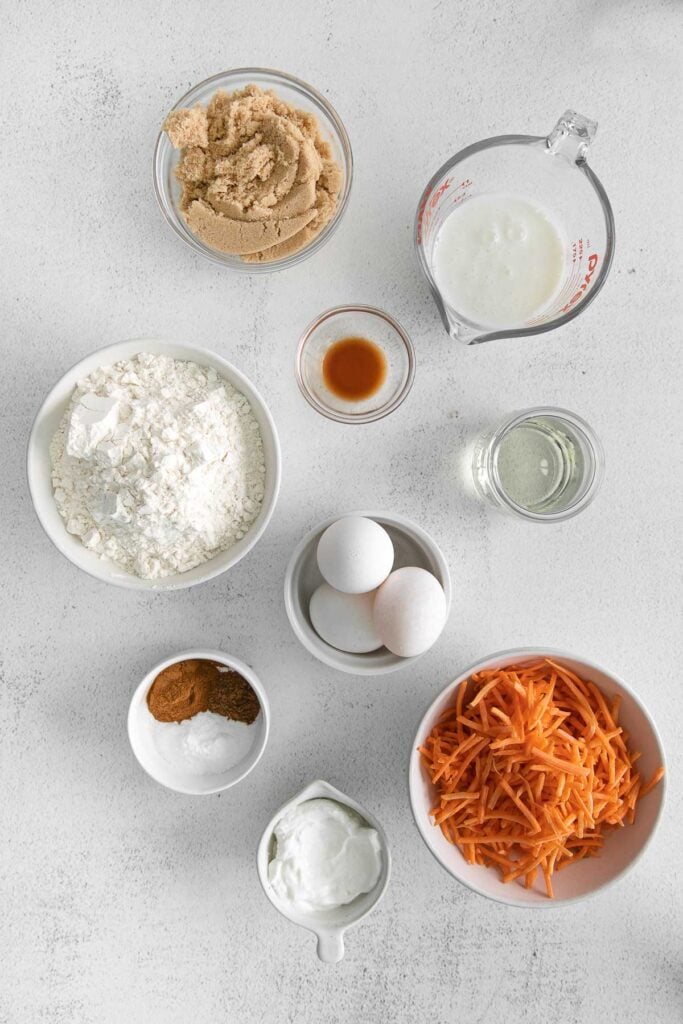 several small white bowls with ingredients for carrot bread: shredded carrots, eggs, flour, yogurt, brown sugar, oil, milk, vanilla yogurt and spices