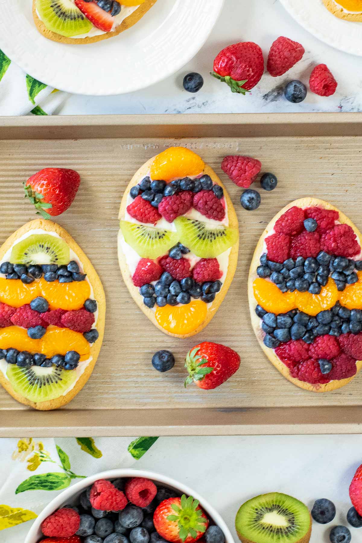three individual sized fruit pizzas topped with strawberries, blueberries, kiwi and oranges on a wood cutting board