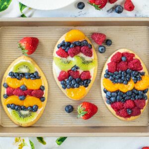 a baking sheet with three egg shaped mini fruit pizzas topped with frosting and fresh berries