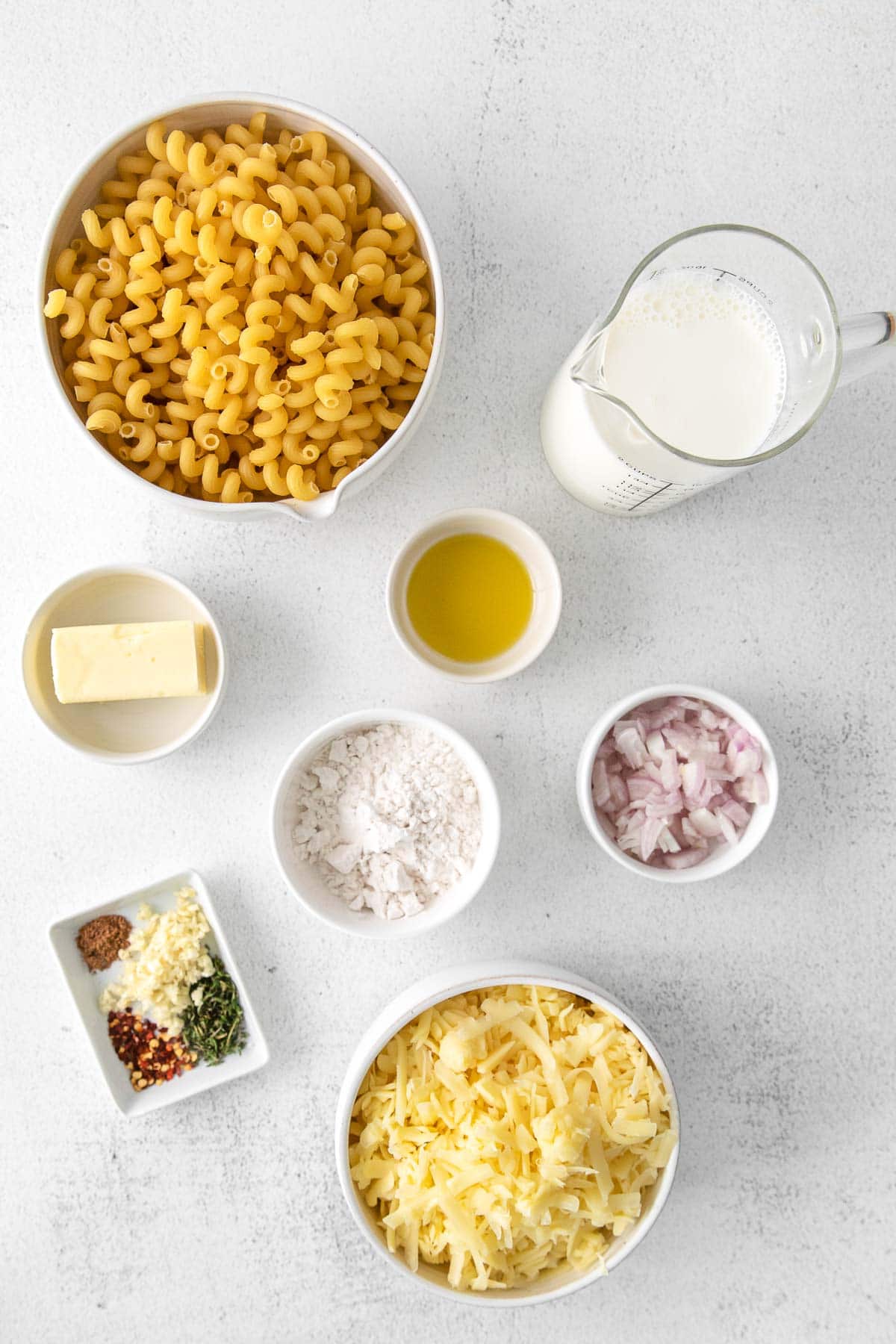 several white bowls with ingredients for homemade mac and cheese - shredded white cheddar cheese, macaroni noodles, milk, butter, flour, onions and spices