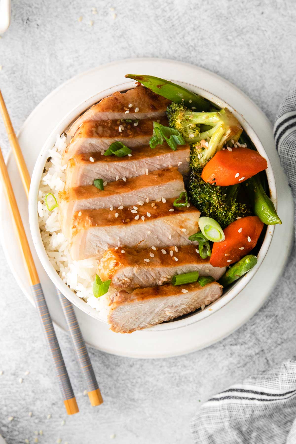 closeup of a white bowl with slices of pork chop, broccoli florets, sliced carrots and green onions with chopsticks on the side