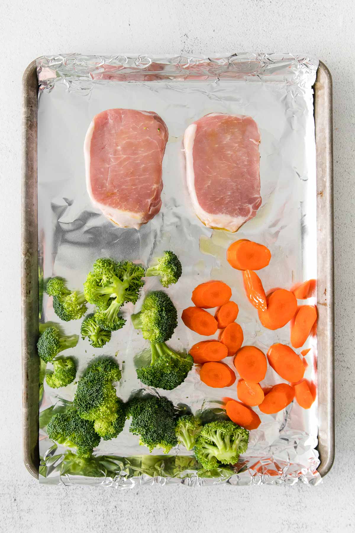 baking sheet with two raw pork chops, broccoli florets and sliced carrots