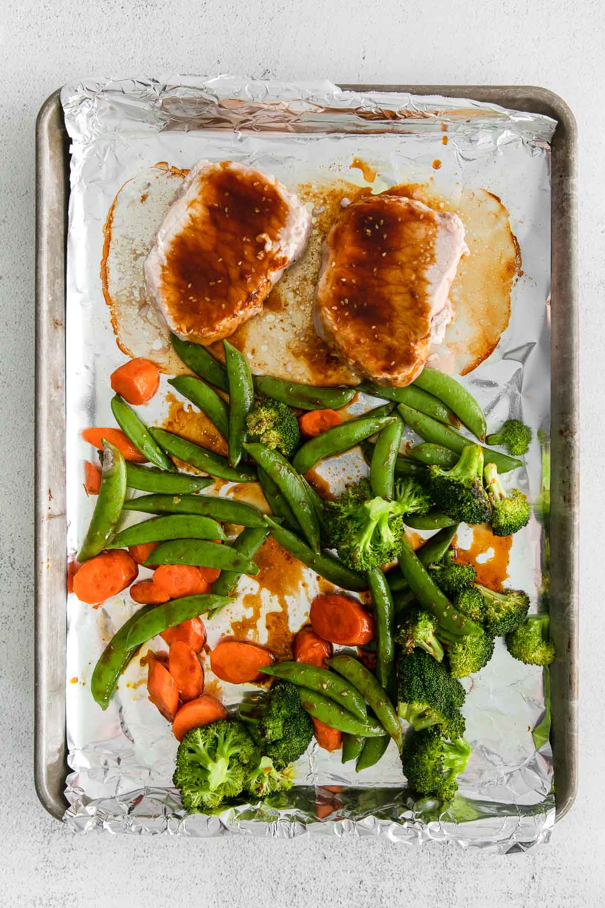 baking sheet with 2 pork chops, carrots, broccoli and sugar snap peas topped with soy marinade