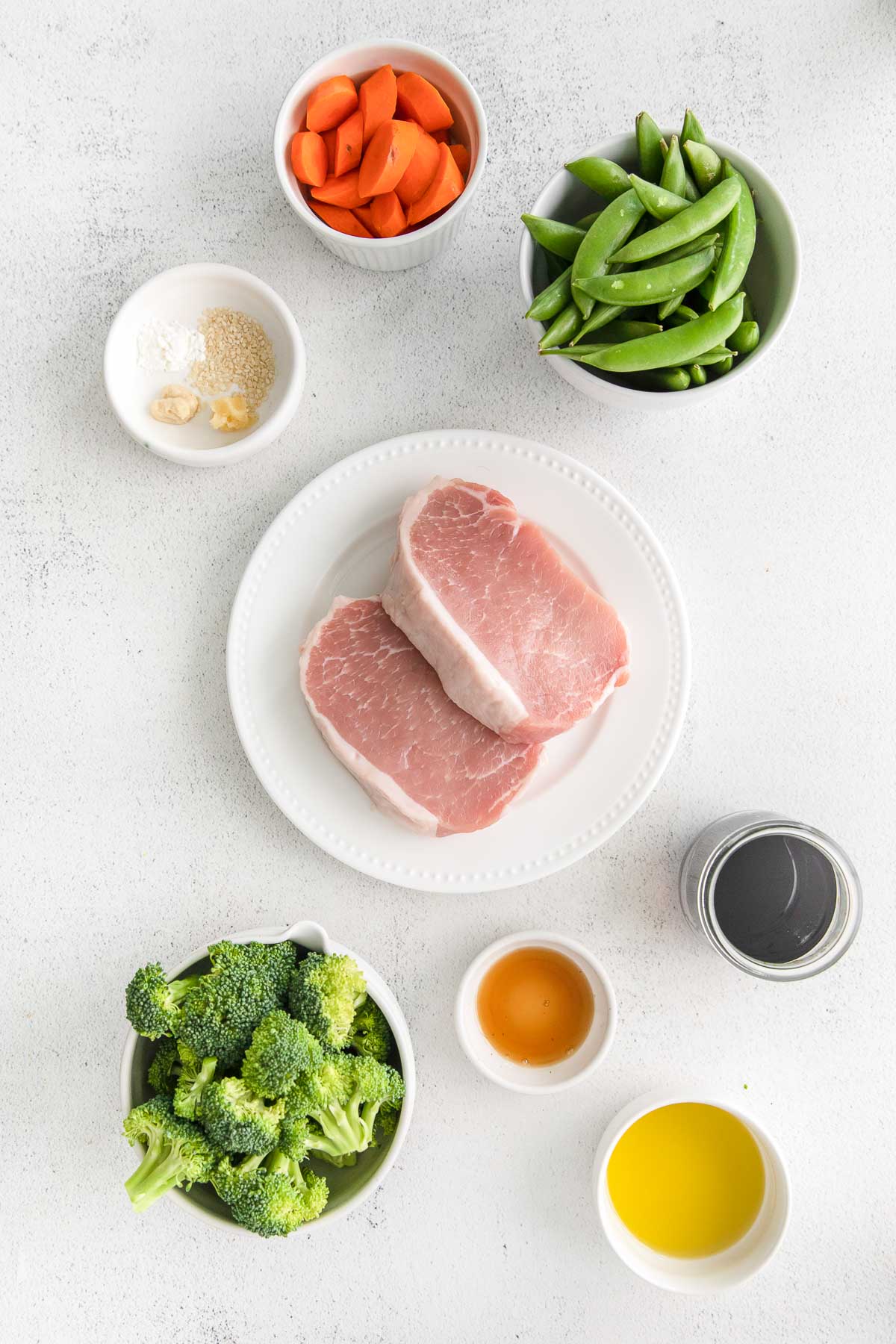 several white bowls with ingredients for asian pork chops - two thick cut pork chops, broccoli florets, snap peas, cut carrots, soy sauce, oil , garlic and ginger
