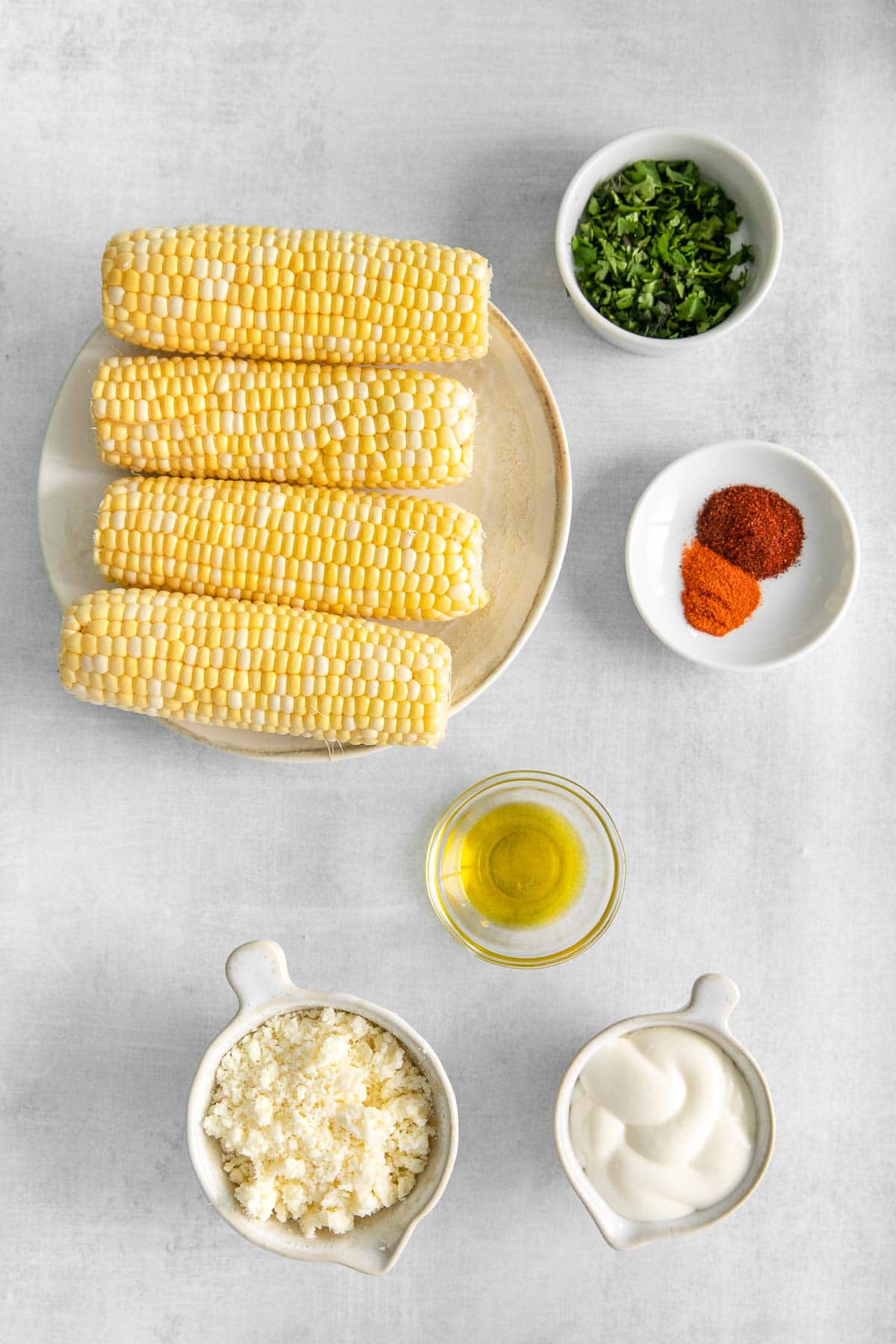several white bowls with ingredients for mexican street corn dip - four corn cobs, crumbled cojito cheese, sour cream, chopped cilantro, olive oil and spices