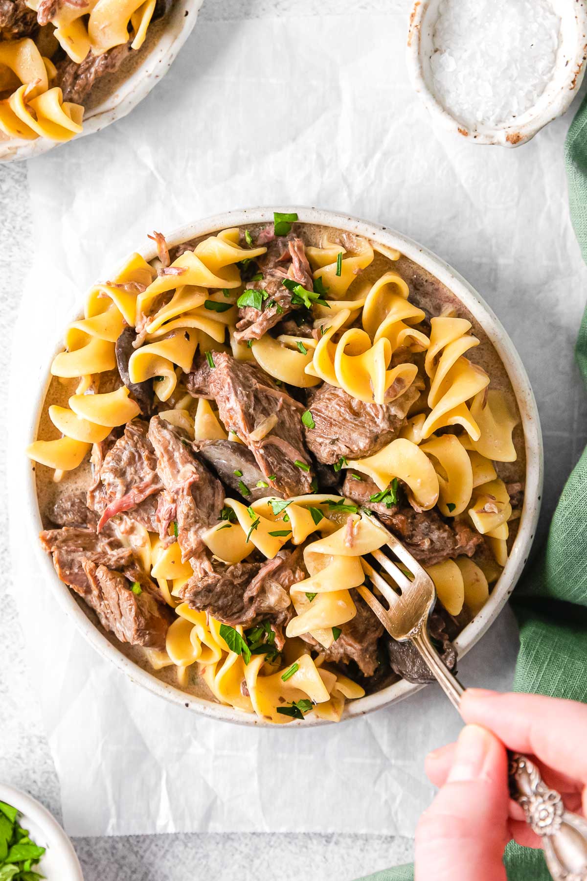 beef chunks and egg noodles in a white bowl with a fork being held by a woman's hand