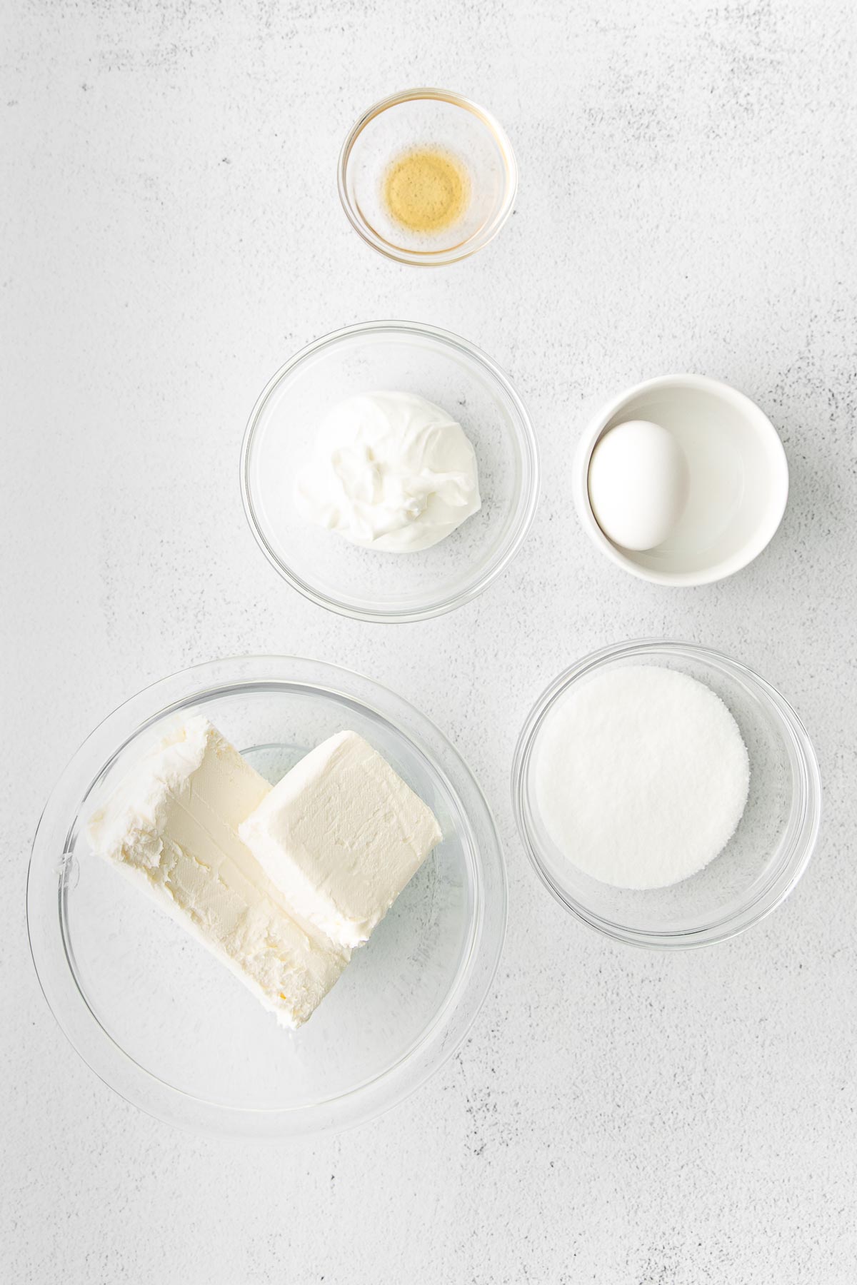 five glass bowls with ingredients for cheesecake filling - cream cheese, sugar, vanilla extract, sour cream and one egg