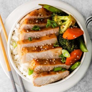 closeup of slices of asian pork chops with broccoli, scallions, sugar snap peas and carrots in a white bowl