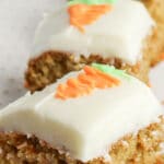three square carrot cake bars topped with cream cheese frosting with an orange icing carrot on top of each