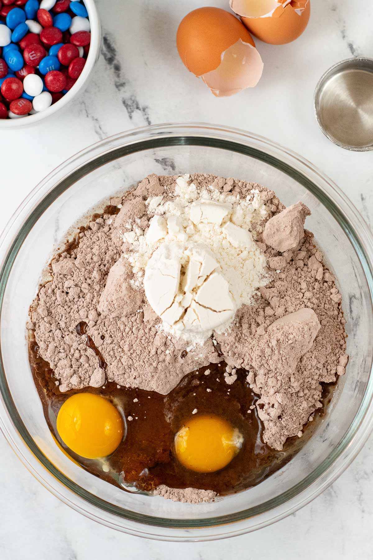 large glass mixing bowl with dry brownie mix, flour and 2 raw eggs