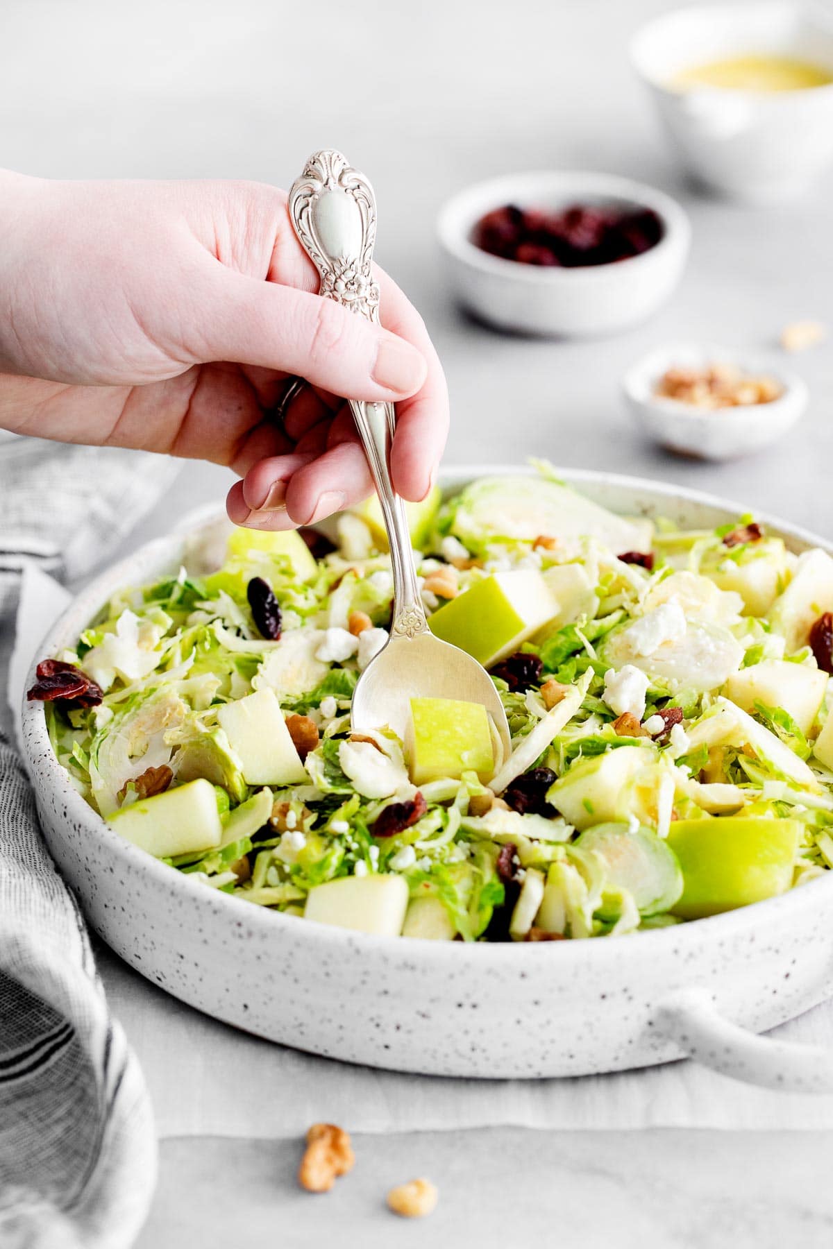 silver spoon scooping brussel sprout salad with chopped green apple out of a white serving bowl.