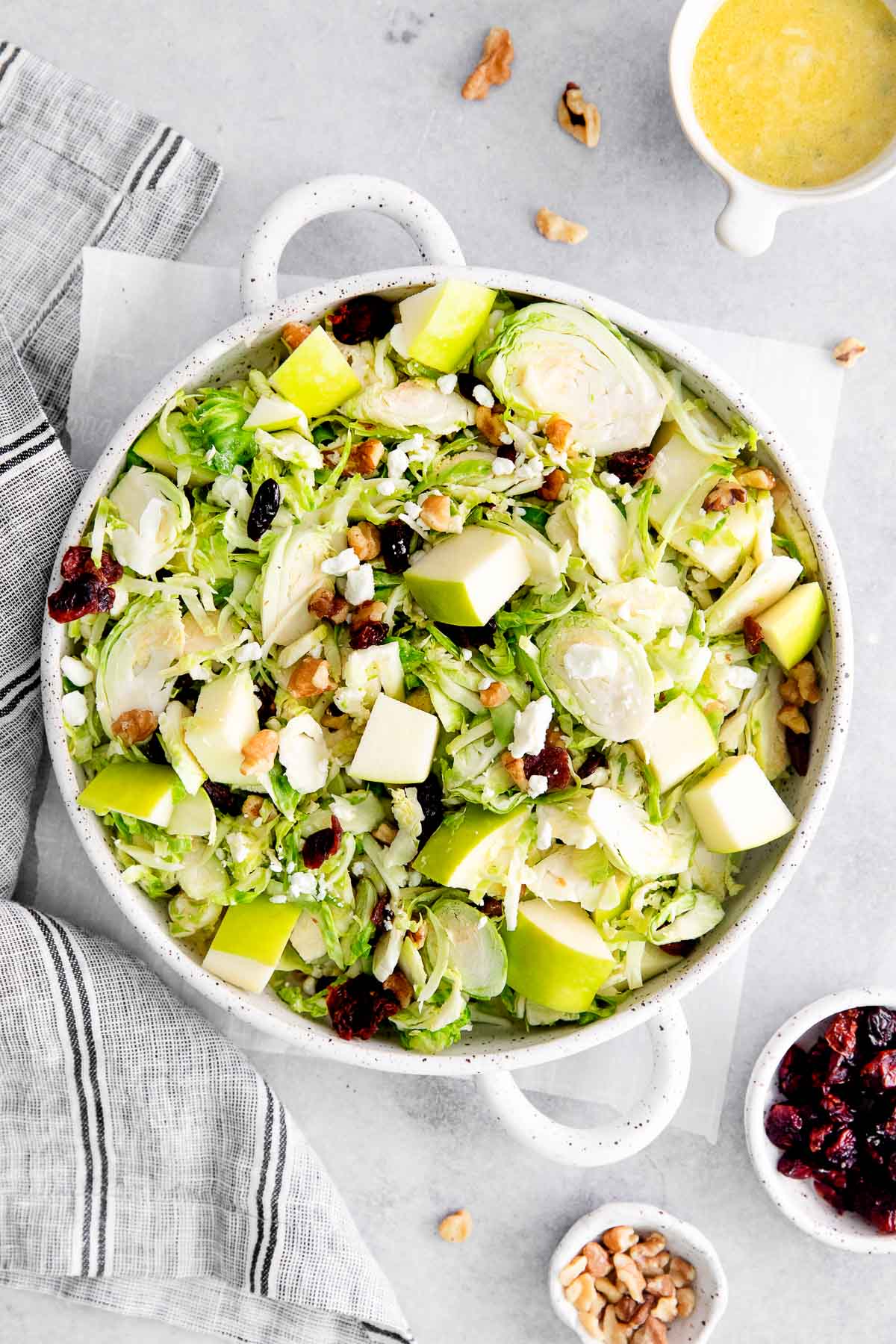 closeup of shaved brussel sprout salad with apples surrounded by bowls of chopped walnuts, dried cranberries, and mustard vinaigrette