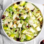 closeup of a white serving bowl full of shaved brussel sprout salad with apples, walnuts and dried cranberries