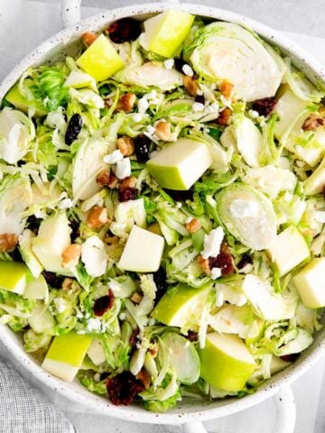 closeup of a white serving bowl full of shaved brussel sprout salad with apples, walnuts and dried cranberries