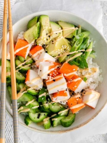 bowl full of fresh cucumber slices, imitaion crab meat, and avocado with spicy mayo drizzles and topped with sesame seeds