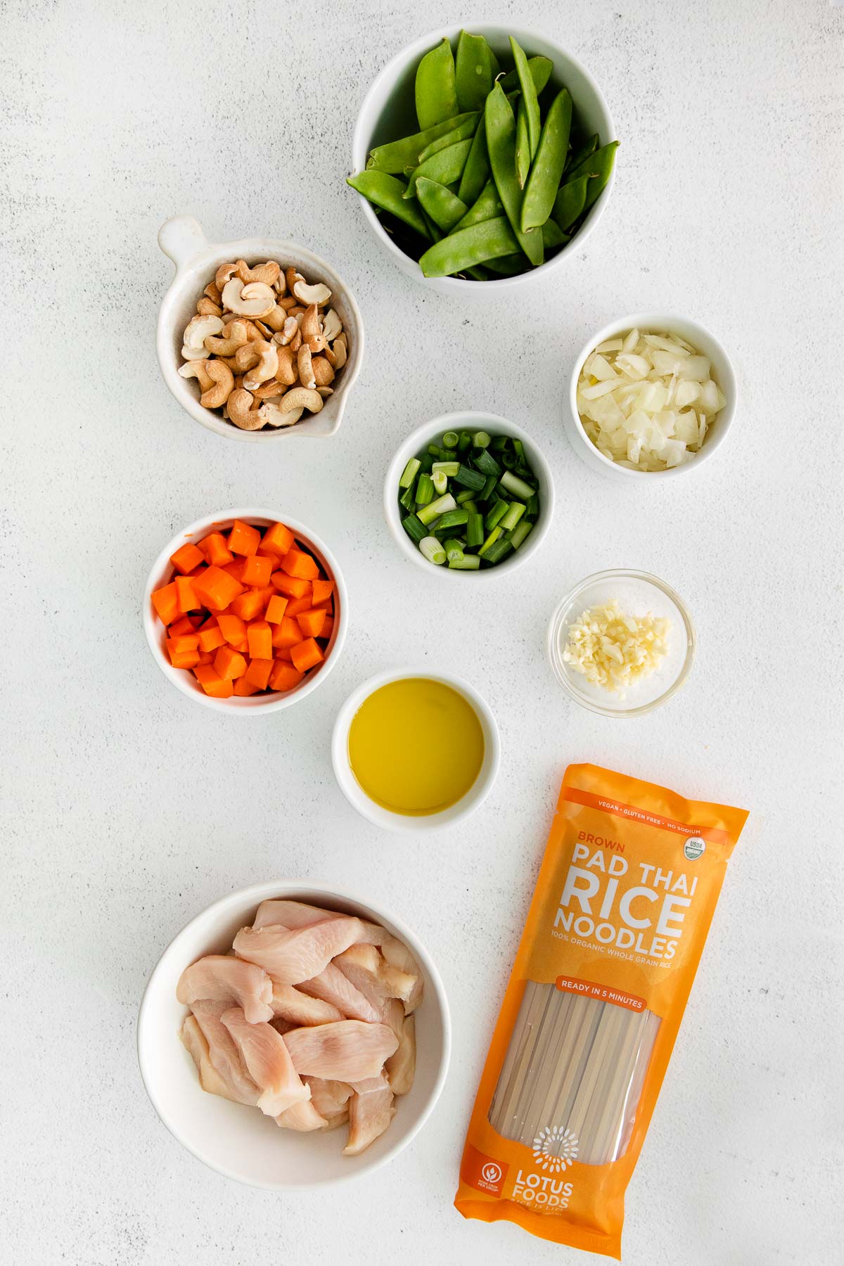 several white bowls with ingredients for cashew chicken stir fry - raw chicken, soy sauce, carrots, snow peas, cashews, garlic, onion and olive oil and a package of lotus foods brown rice noodles