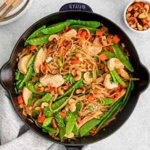 closeup of cast iron skillet of thai cashew chicken stir fry with rice noodles and snow peas and sesame seeds