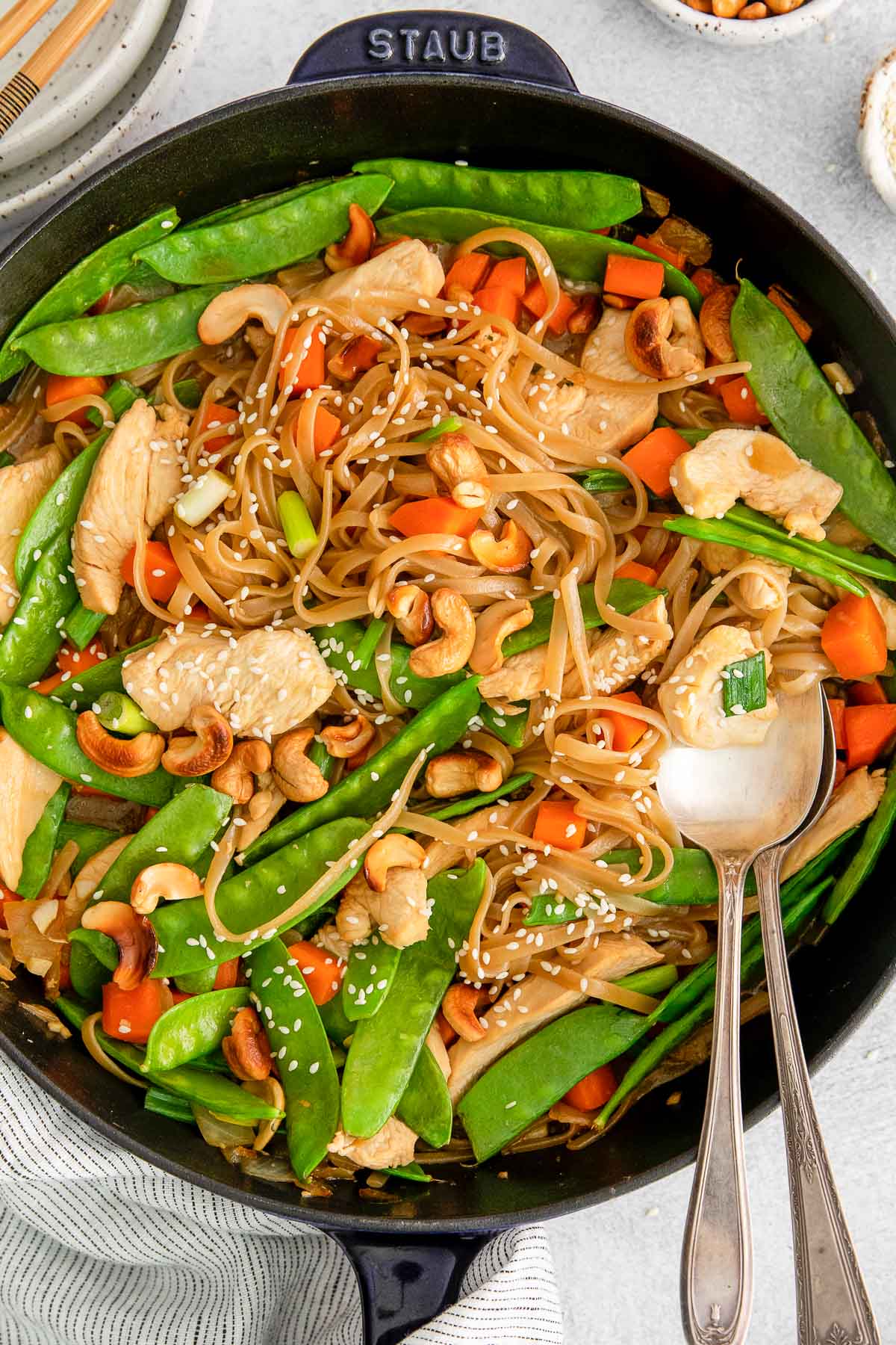 closeup of stir fry with chicken, cashews, snow peas, carrots and rice noodles in a brown sauce with two silver spoons