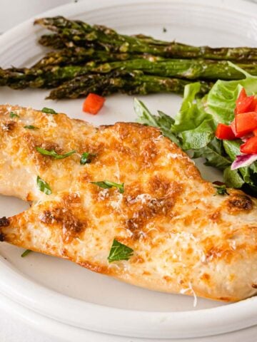 parmesan crusted chicken breast on a white plate with asparagus spears and salad