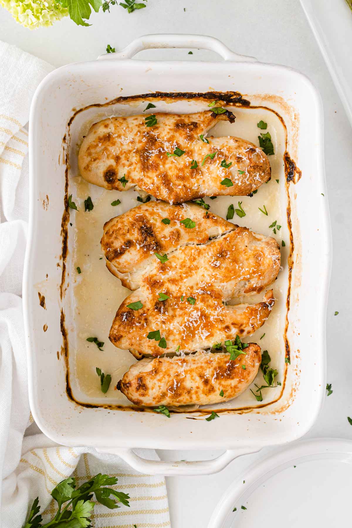 white casserole dish with four baked chicken breasts topped with parmesan cheese and fresh parsley