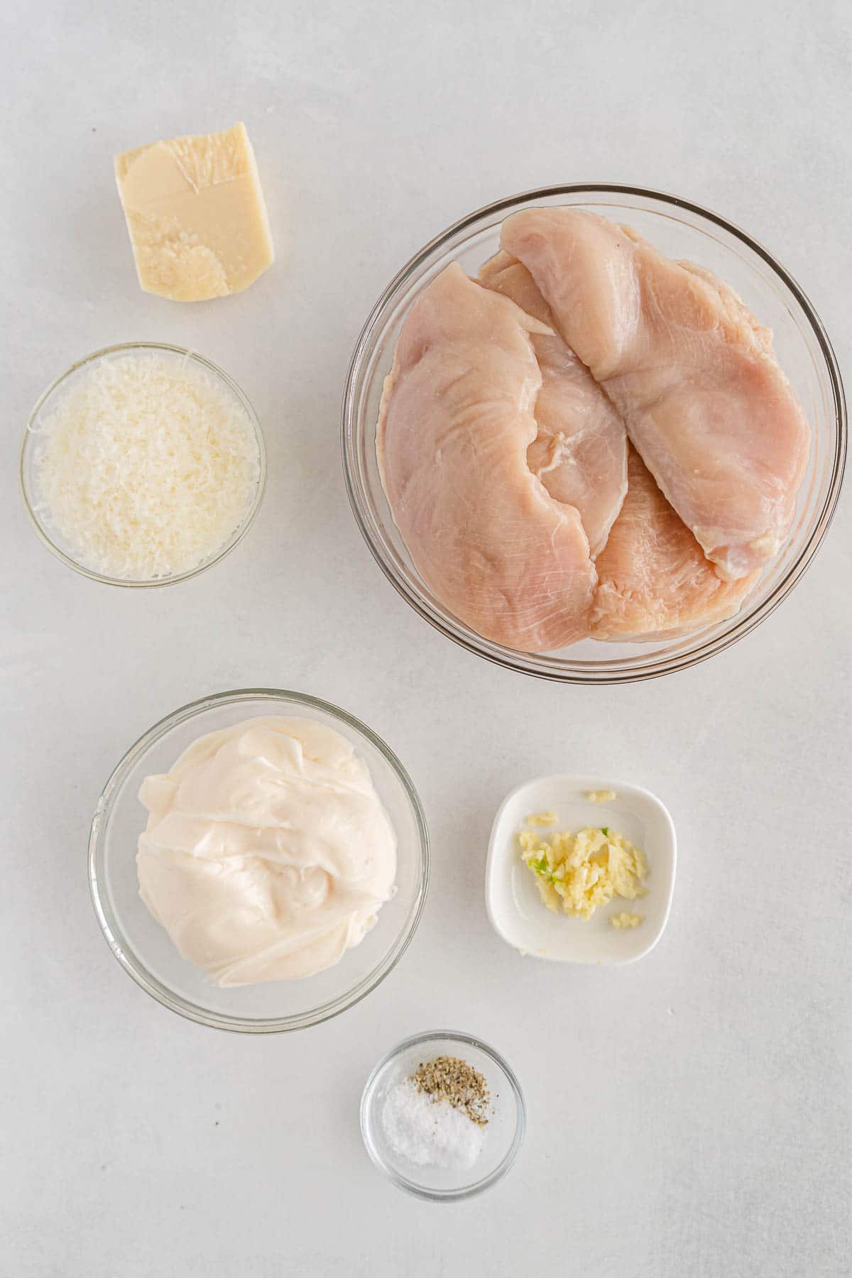 several small glass bowls with ingredients for parmesan chicken - raw chicken breasts, mayonnaise, parmesan cheese, minced garlic and salt and pepper