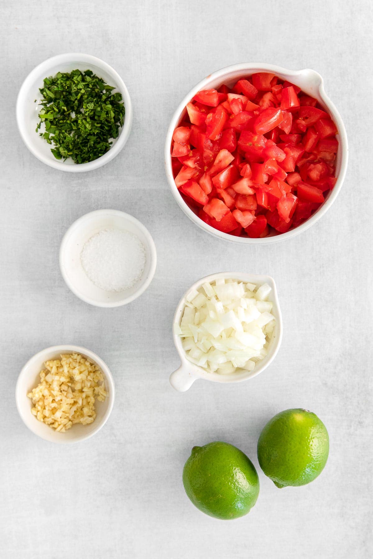 several white bowls with ingredients for pico de gallo - diced tomatoes, diced onions, minced garlic, two limes. and fresh chopped cilantro