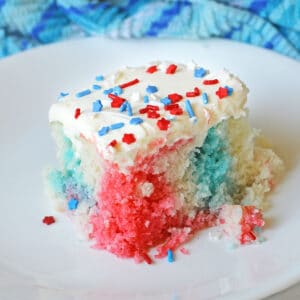 slice of red white and blue poke cake with with white icing and 4th of July sprinkles on a white plate