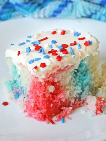 slice of red white and blue poke cake with with white icing and 4th of July sprinkles on a white plate