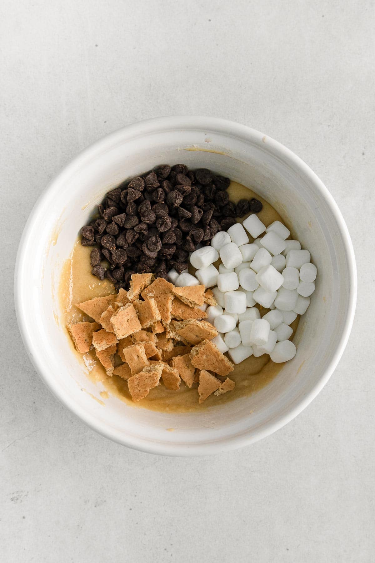 white mixing bowl with cookie batter topped with mini marshmallows, chocolate chips, and graham crackers chunks