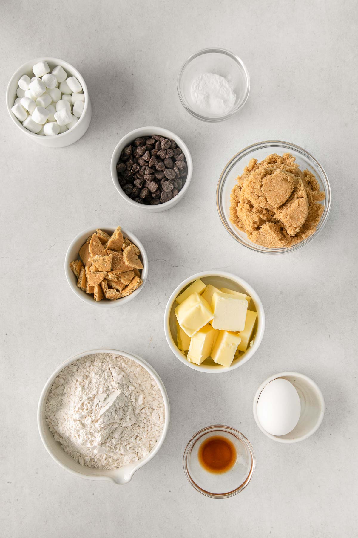 several white bowls with ingredients for s'mores cookies - flour, brown sugar, butter, vanilla, egg, mini marshmallows, chocolate chips, graham crackers, baking powder
