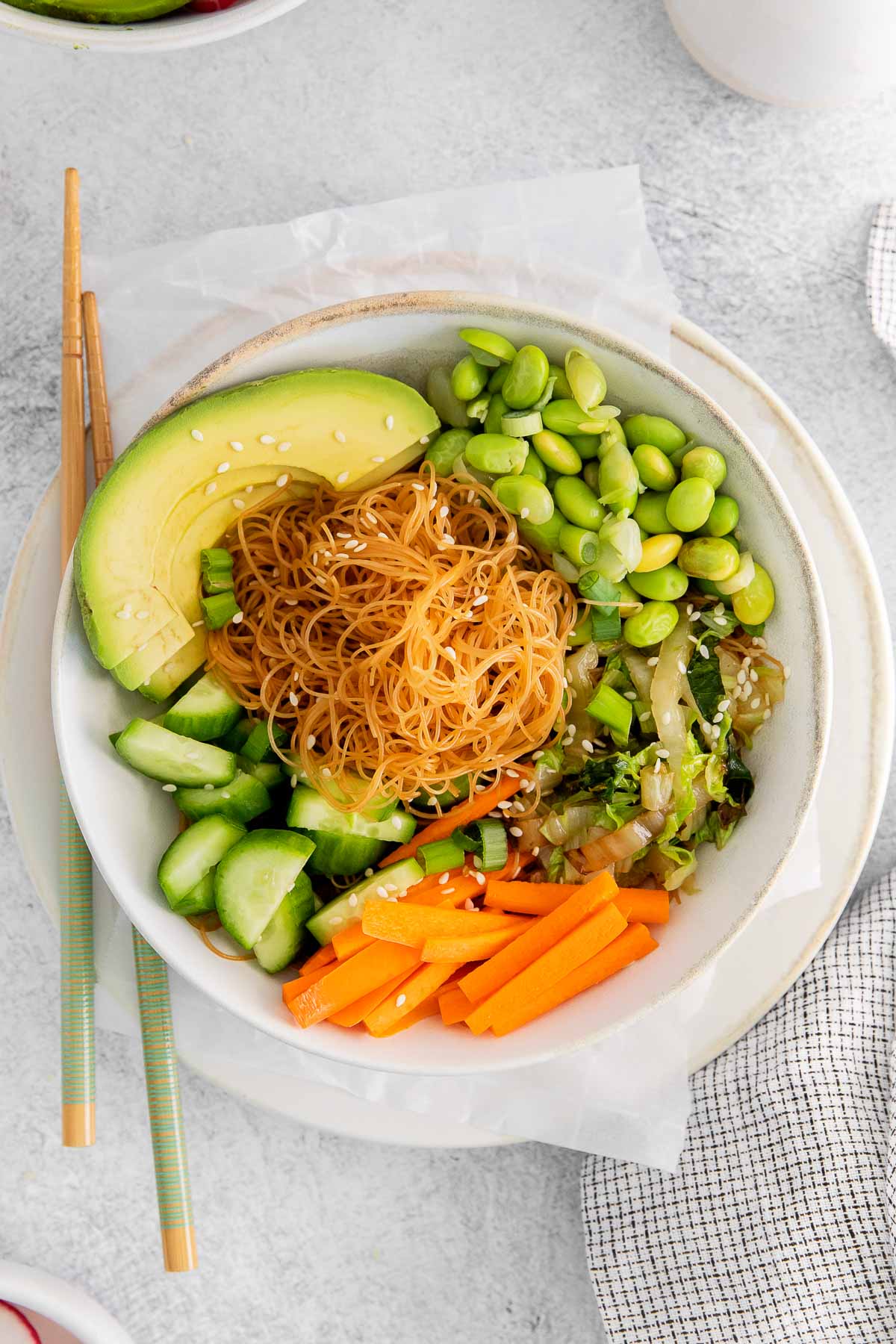 white bowl with sliced carrots, edamame, diced cucumbers, rice noodles and avocado topped with sesame seeds with chopsticks on the side of the bowl