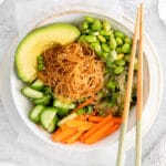 Close up of a white bowl with rice noodles surrounded by edamame, sliced carrots, cucumbers, and avocado topped with sesame seeds